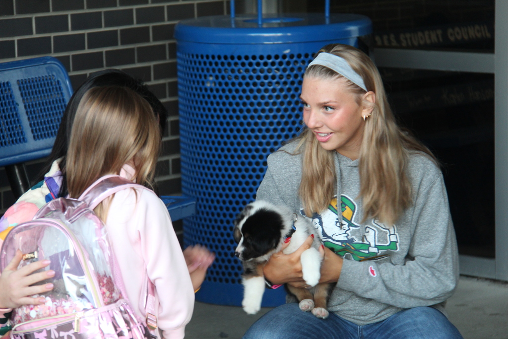 One of the athletes puts her arm around one of the elementary students as they look at the puppy in the arms of another athlete across from them. 