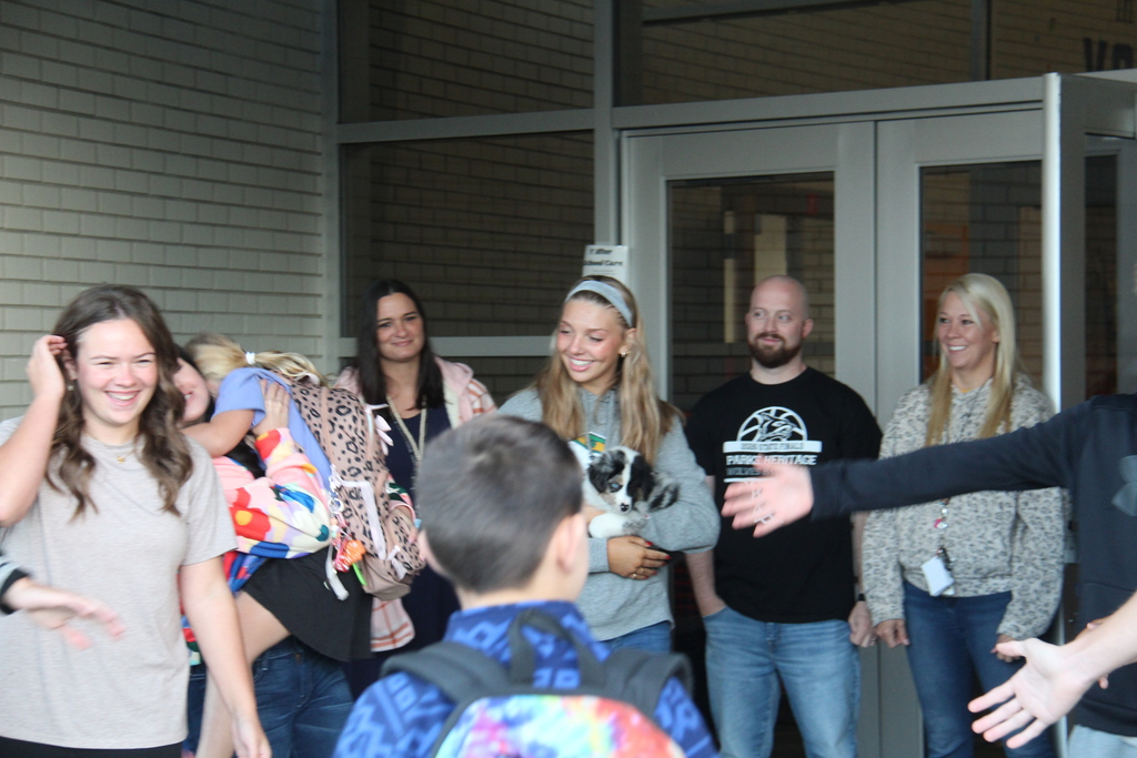 Members of the Tennis, Cross Country, and Golf teams line up along the sidewalk to five high fives to one of the students as he walks into school. Another student in the background hugs one of the athletes in the background. 
