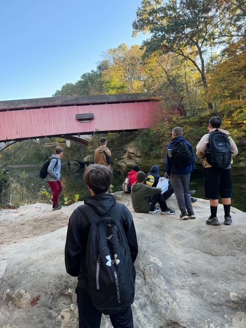 Photo of kids standing on a large rock, facing the Narrows Covered Bridge. 