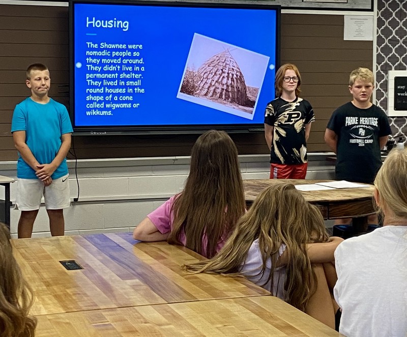 three students standing by a computer monitor talking about Native Americans