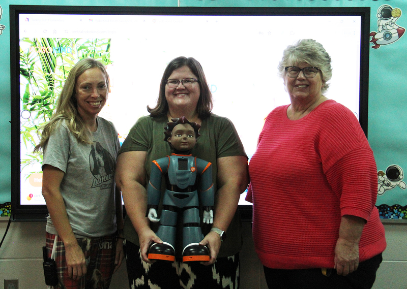 three women standing in front of a smartboard with one of them holding a robot