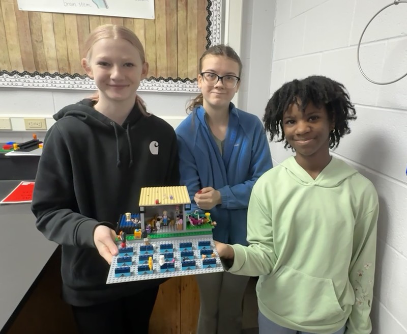 three students standing together holding a theater set made out of Legos