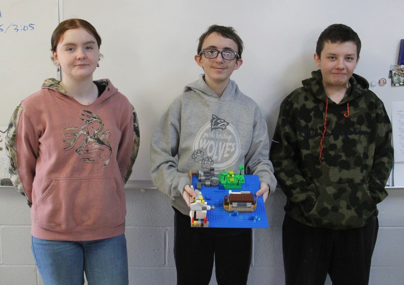 three students standing in a line with the one in the middle holding a theater set made out of Legos