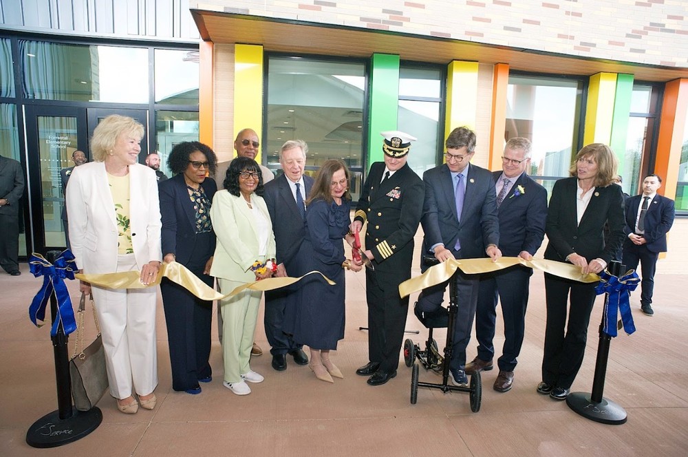 A photo of nine individuals cutting a long golden ribbon to dedicate the new Forrestal Elementary School at Naval Station Great Lakes. Included in the lineup, from left to right, are North Chicago Mayor Leon Rockingham, a woman in a suit, a woman in a dark patterned blazer, Dr. John Price (Superintendent of North Chicago School District 187), Lake County Regional Superintendent of Schools Lori Thomas, Captain Stephen Yargosz (Commanding Officer Naval Station Great Lakes), U.S. Congressman Brad Schneider (who uses a knee scooter), Illinois State Senator Adriane Johnson, and an unidentified man. Behind them, the school building features colorful vertical facade panels in green, orange, and yellow.