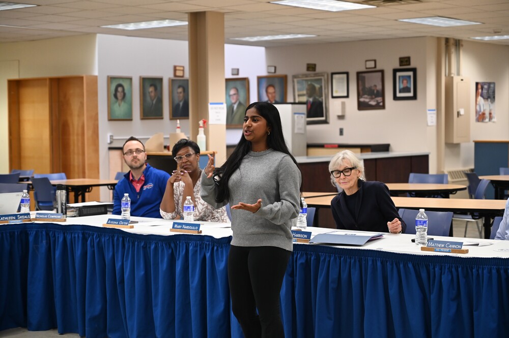 A student speaks at the front of the room as members of the Board of Education look on, seated behind her.