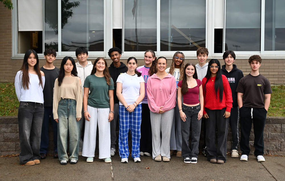 15 students pose in front of a low cement wall and building after being selected for the NYSSMA All-State Festival.