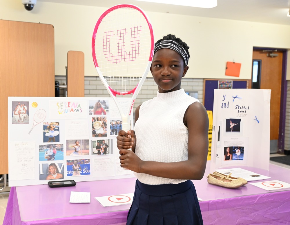 A student holding a tennis racquet presents her project on Serena Williams.