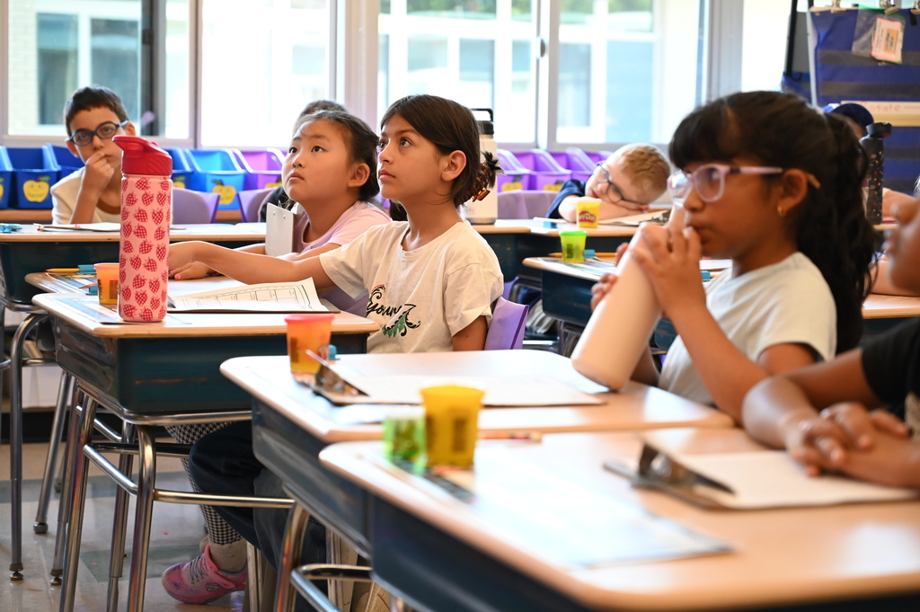 Students listen intently from their desks as their teacher reads at the front of the room.