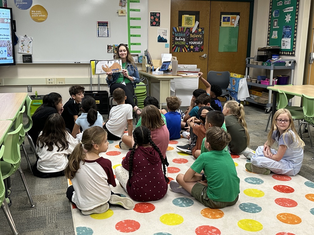 The librarian reads a book to a class of students who sit on a carpet in front of her.