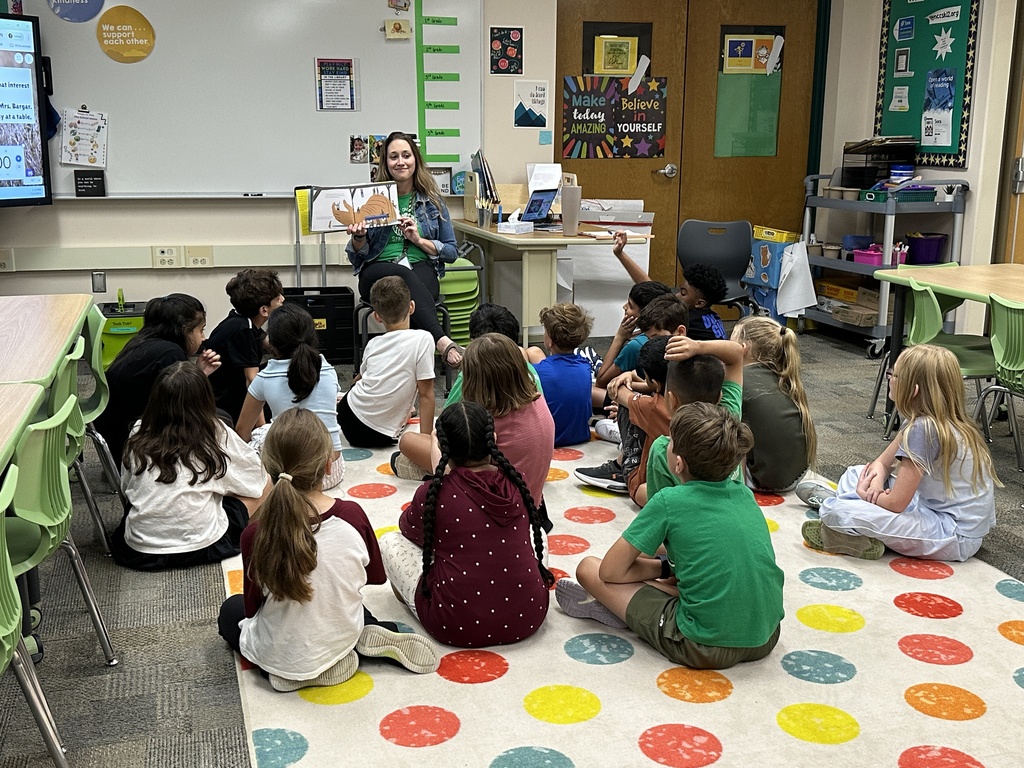 Students look on from the carpet as the librarian reads them a book.