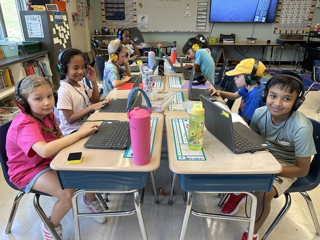 A group of students wearing headphones sit at their desks and work on their Chromebooks.