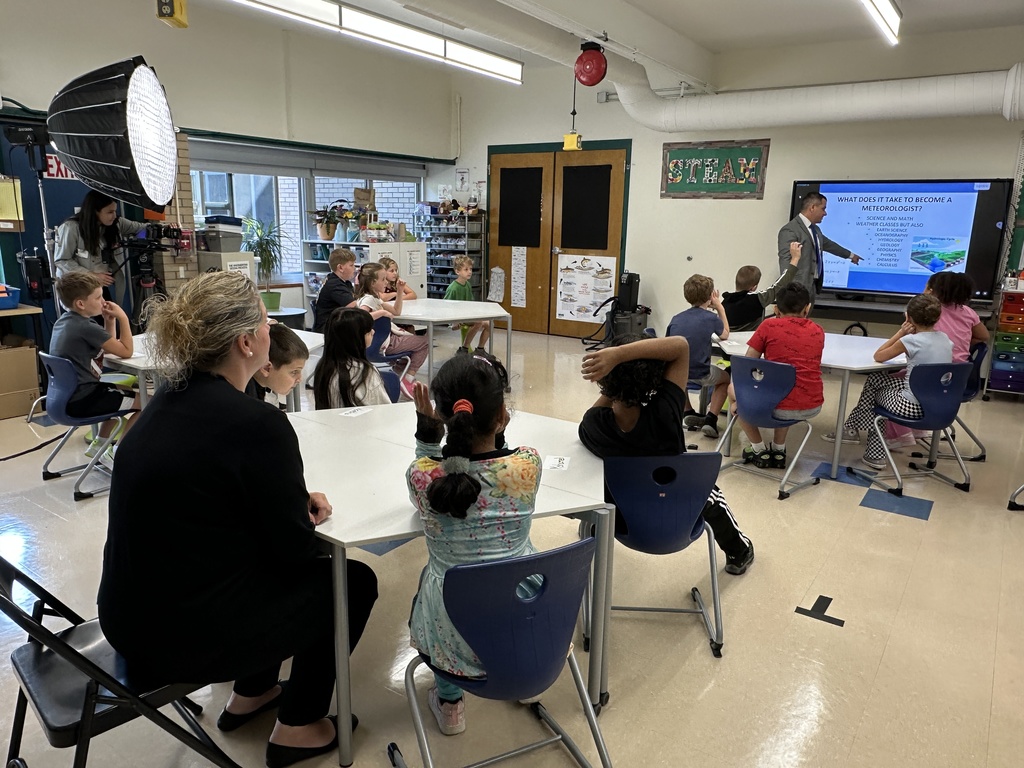 Meteorologist Chris Gilson stands at the front of a room of students and points to a slideshow presentation on weather.