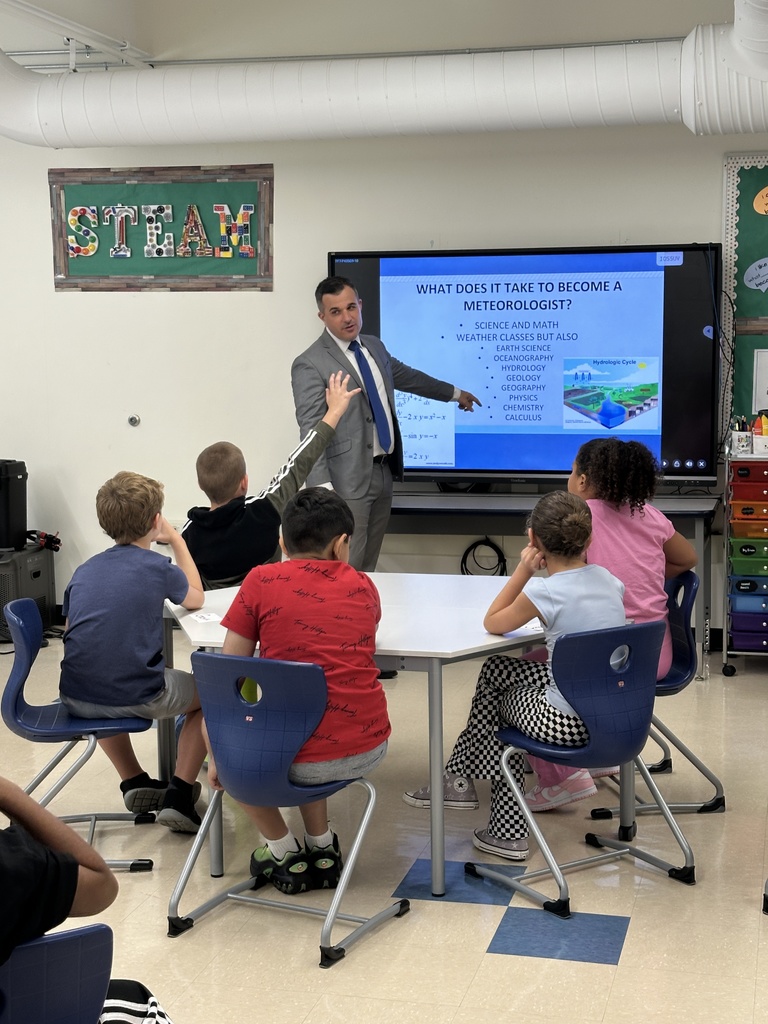 Meteorologist Chris Gilson stands at the front of a room of students and points to a slideshow presentation on weather.
