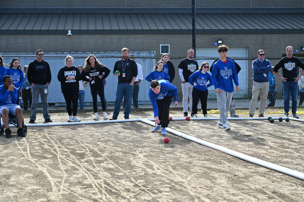 A player rolls the bocce ball down the court as teammates and opponents look on behind him.