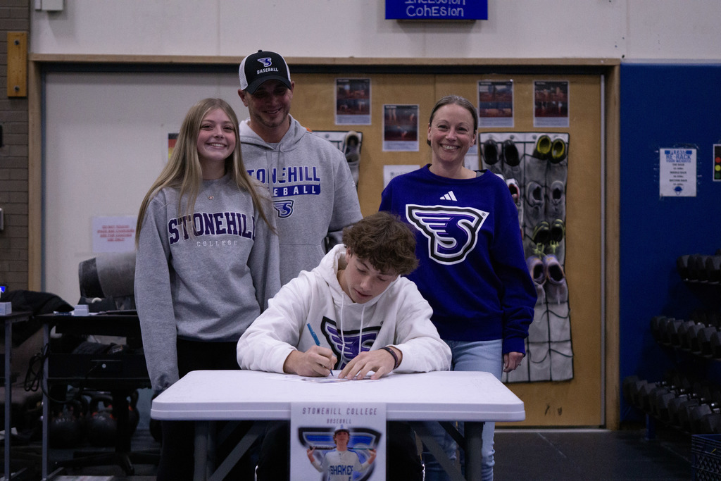 A student signs his letter of intent surrounded by his family members looking on.