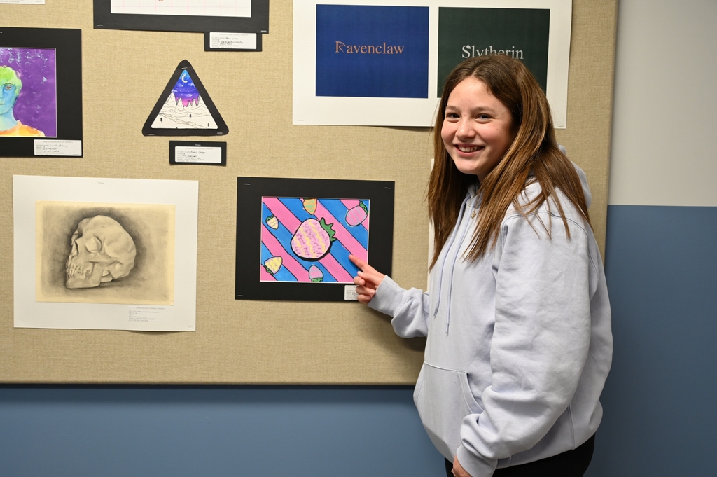 A student proudly points to her work of art displayed in a gallery.