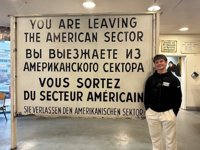 you are leaving the american sector sign, with text below in different languages. student is standing in front of the sign