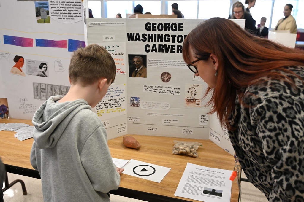 A student reads his presentation on George Washington Carver to a teacher.