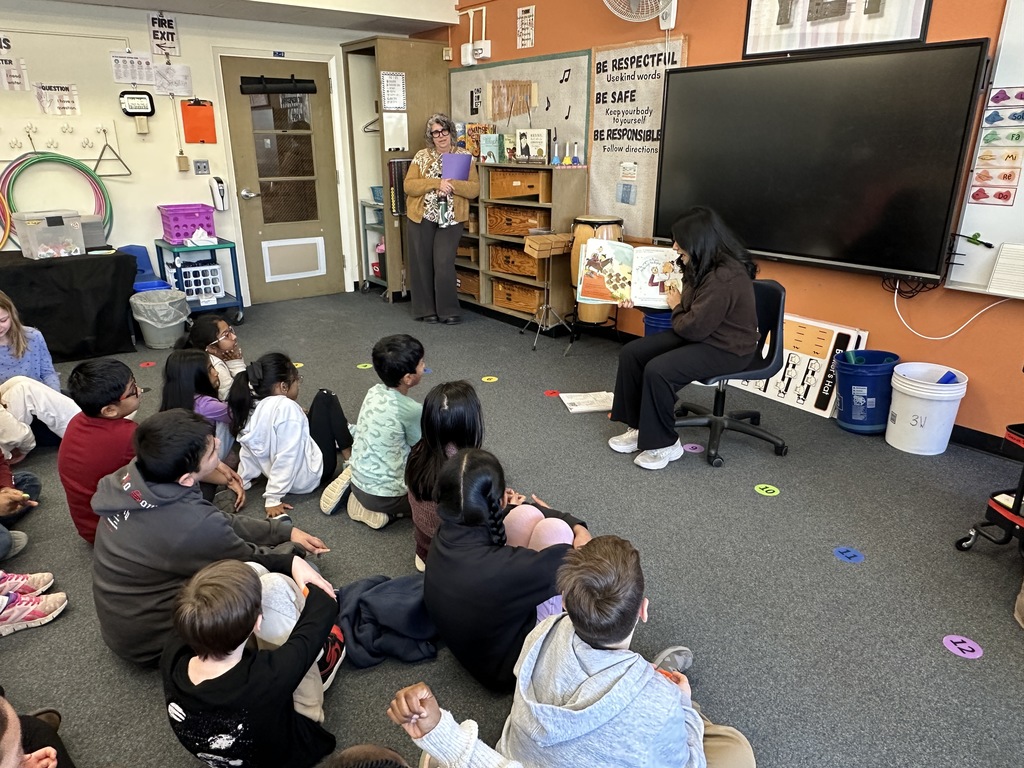 A high school student sits in a chair and reads a book to a group of elementary school students seated in front of her.