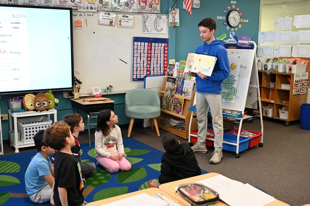 A high school student stands and reads a book to a group of elementary students.