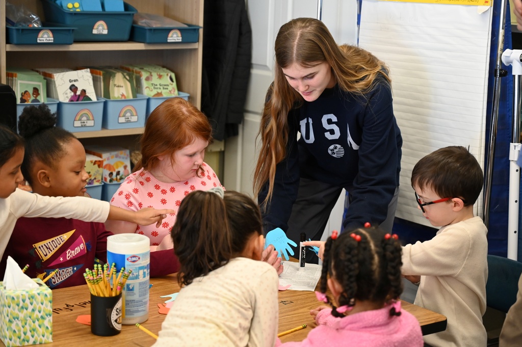 A high school student leads a craft with a group of elementary students at a table.