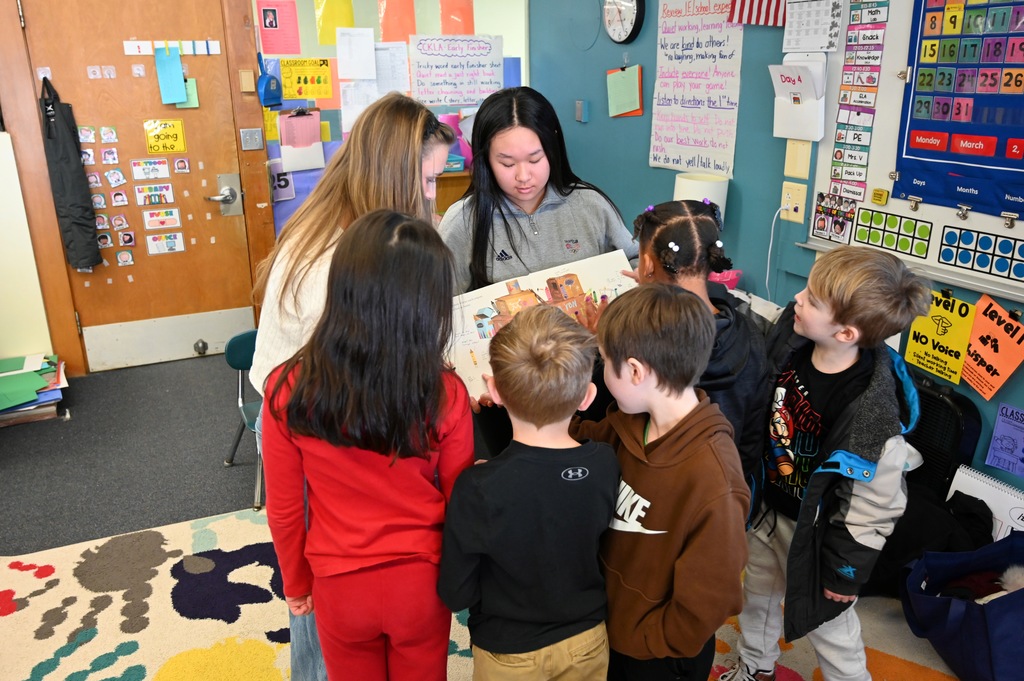 Younger students crowd around two high schoolers as they read a book.