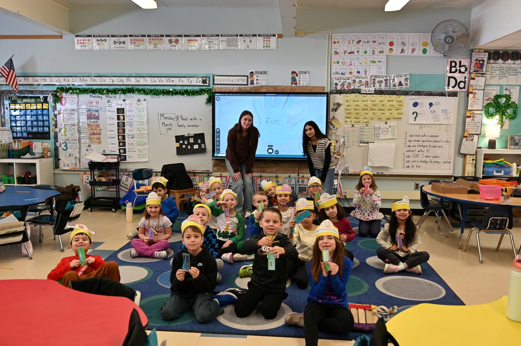 A group of first graders pose with their handmade hats while sitting on the carpet.
