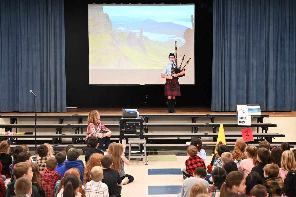 A student stands on stage and plays the bagpipes for a room full of students sitting on the floor.