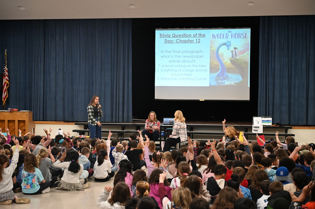 Students sit on the floor and raise their hands to answer trivia questions about the book they're currently reading.