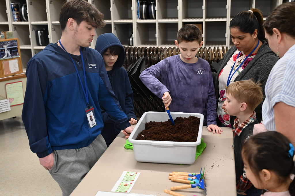 A student digs with a hand shovel as other students and adults stand around watching.