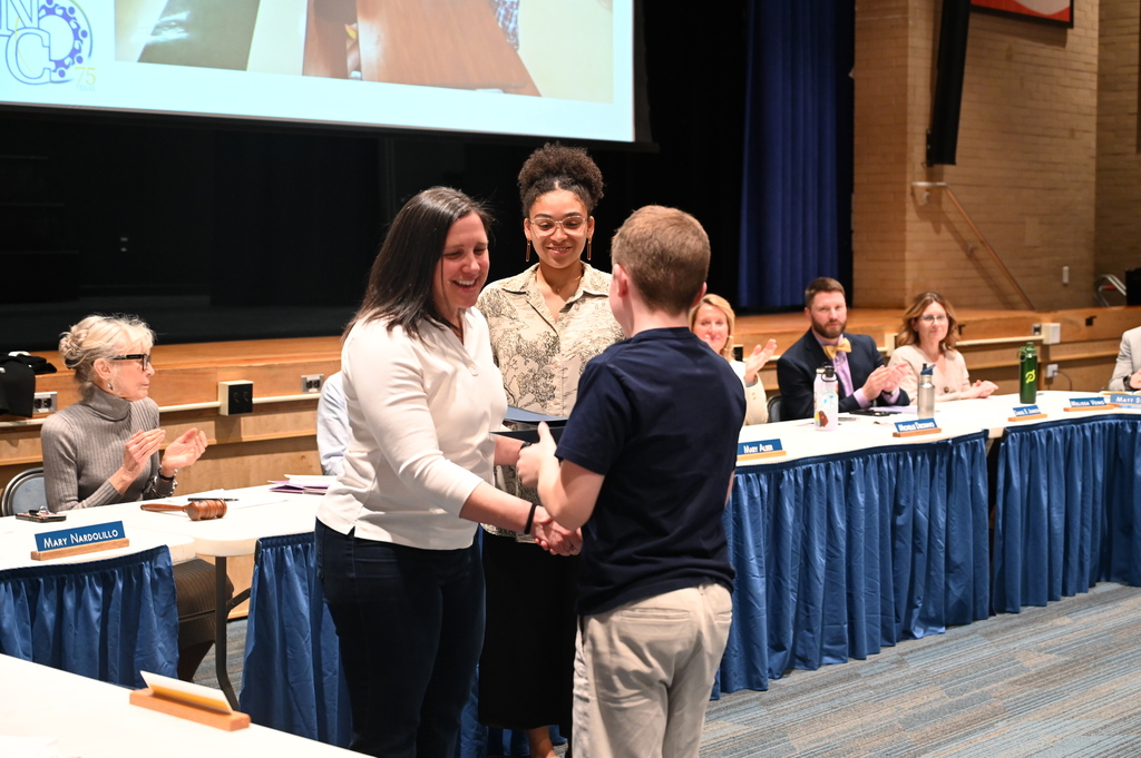 A student receives a certificate from two teachers as the members of the Board of Education look on.
