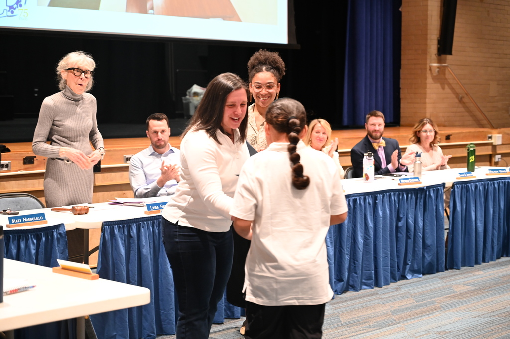 A student receives a certificate from two teachers as the members of the Board of Education look on.