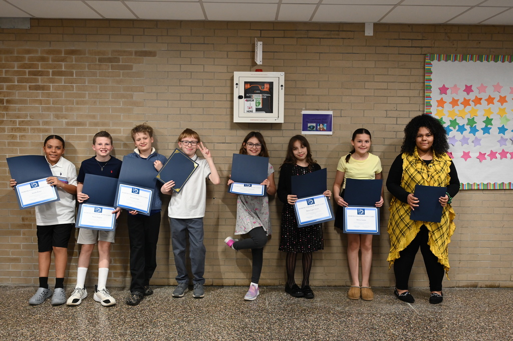 A group of eight students pose against a brick wall with their certificates of recognition.