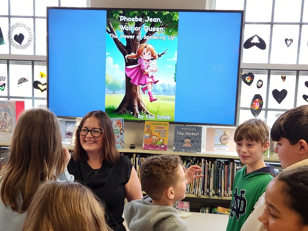 Mrs. Smith smiles as she stands in front of a monitor with one of her book covers on it.