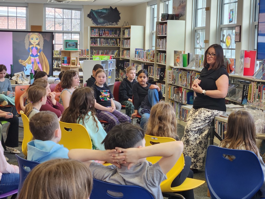 Mrs. Smith stands at the front of the library and speaks to students seated in chairs in front of her.