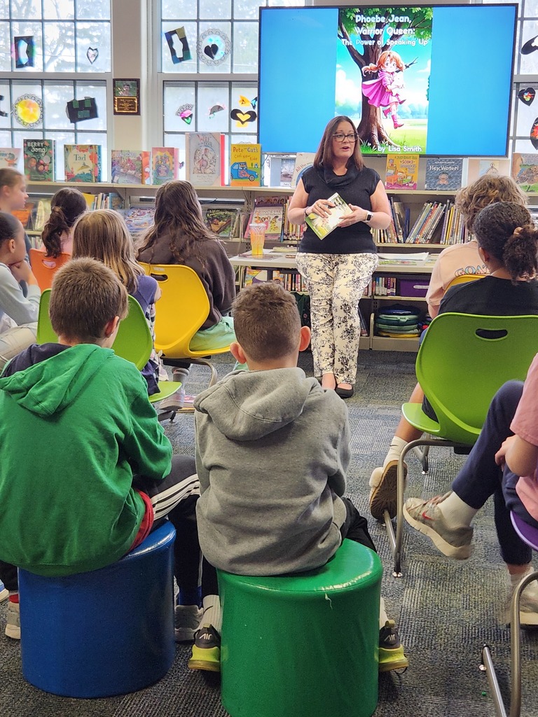 Mrs. Smith holds one of her books in her hands as she speaks to a library full of students.