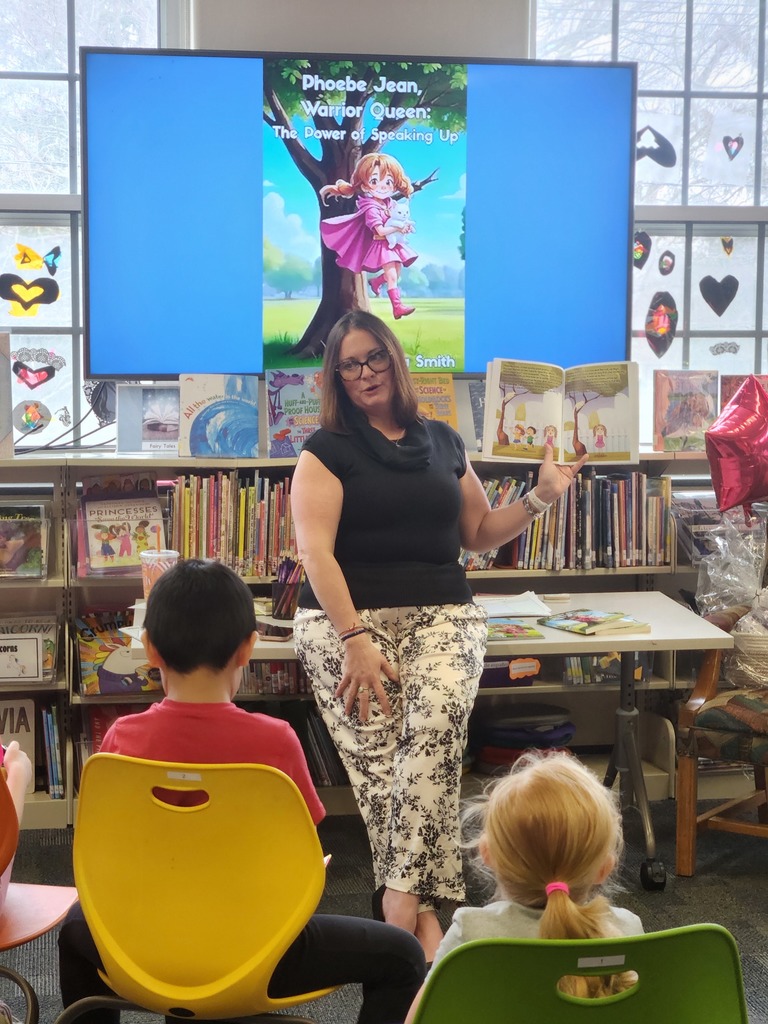 Mrs. Smith holds a book open as she reads it to students seated in front of her.