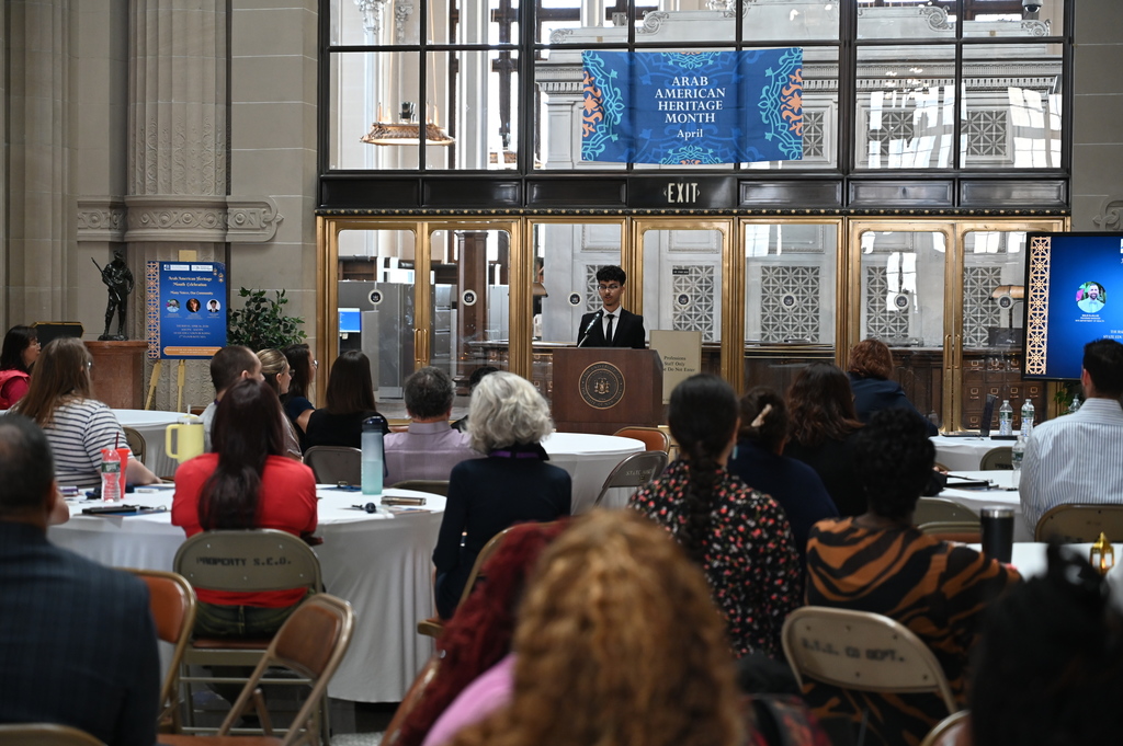 Shaker High School senior Nawaf Kassem stands at a podium, reading a proclamation in front of a room full of people.