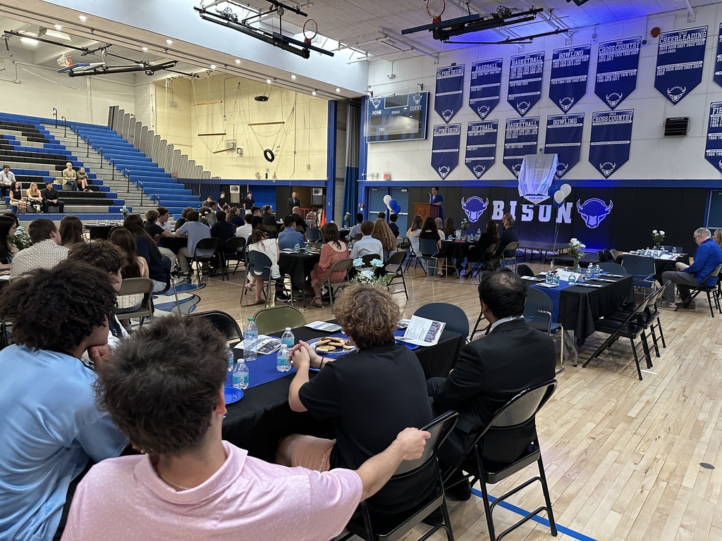 People sit at tables all over a decorated high school gymnasium as a speaker stands at the podium.
