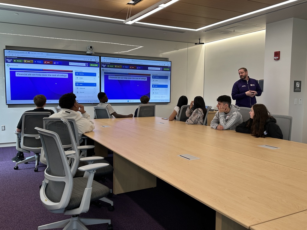 A representative from UAlbany speaks to Shaker students, using questions on two screens as prompts.