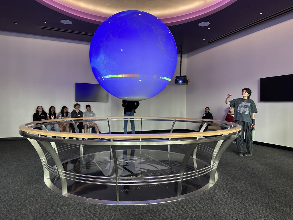 A UAlbany student speaks to high schoolers about atmospheric science, utilizing a large suspended globe to show trends.
