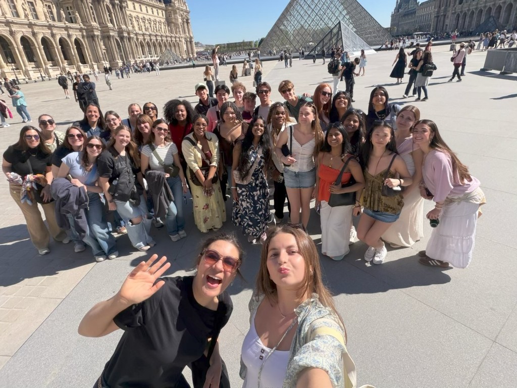 Two people stand in front to take a selfie with a large group of students behind them.