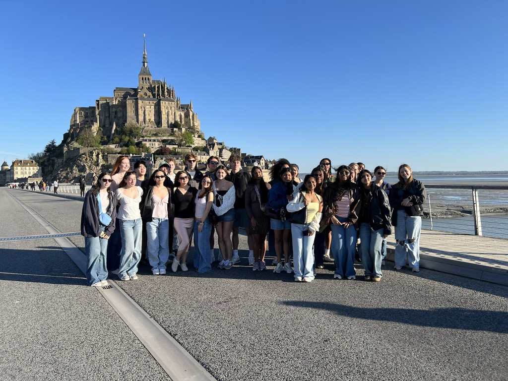 A large group of students stand with a beautiful building and a body of water behind them.