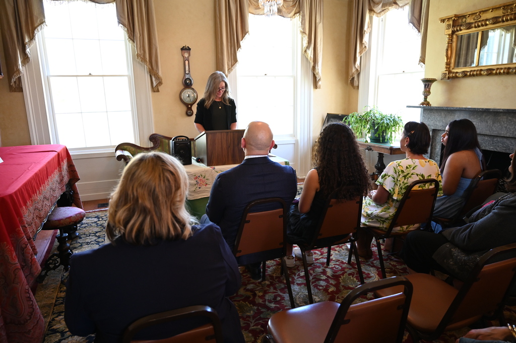 A woman stands and speaks at a podium in the front of a room as people look on while seated.