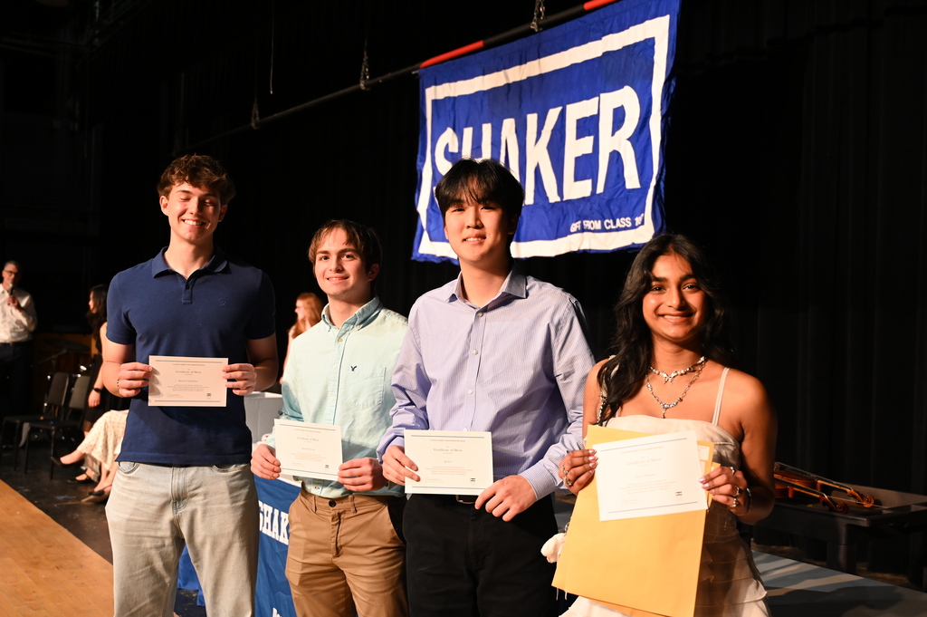 Four National Merit Scholarship Finalists hold their certificates on stage in front of a Shaker backdrop.