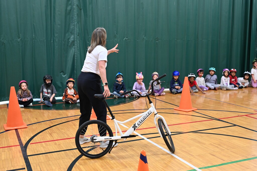 An instructor holding a bike speaks to kids in helmets seated on the gym floor.