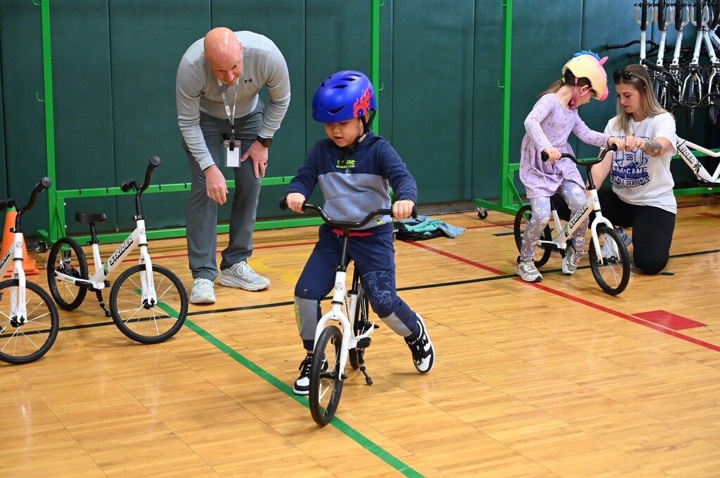 P.E. teachers look on and assist young students learning to ride bicycles in class.