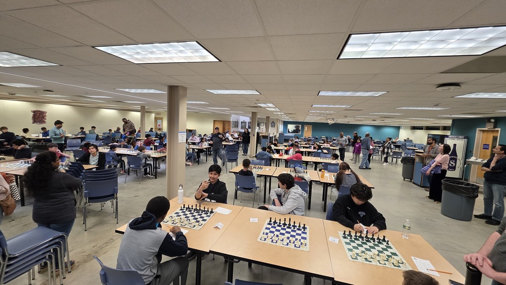 A cafeteria filled with students playing chess as adults look on.