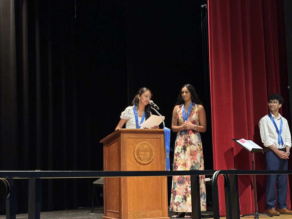 A student speaks into the microphone at the podium on stage as two others look on.