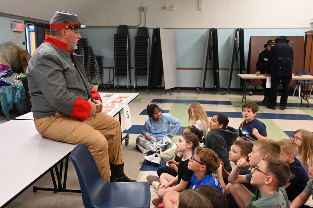 An actor portraying a soldier sits on a table and speaks to a group of students.